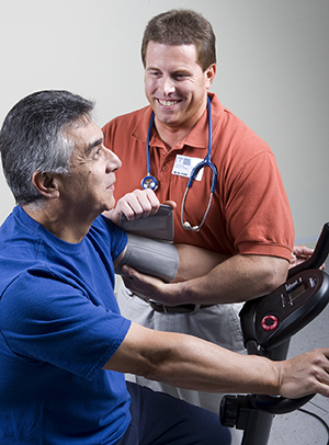 Hombre ejercitando en una bicicleta fija mientras un fisioterapeuta mide su presión arterial.